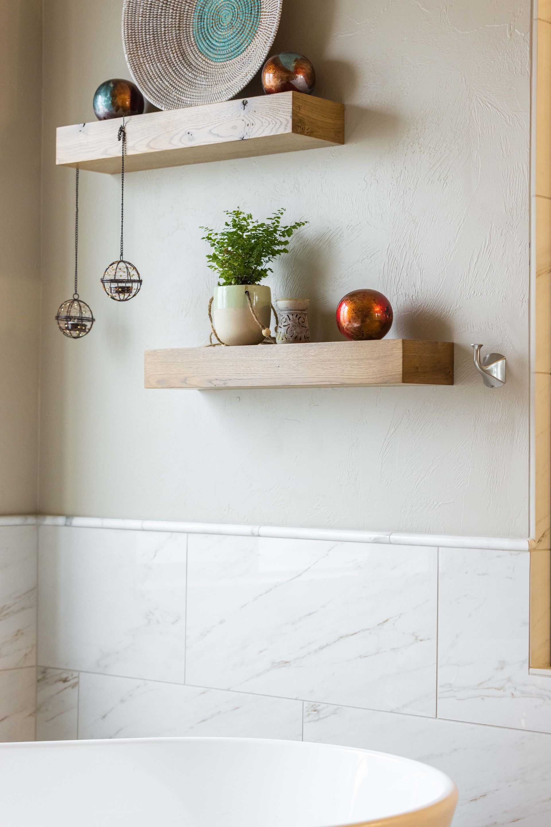 Minimal bathroom with marble tile, floating wooden shelves, a plant, and hanging glass ornaments above a tub
