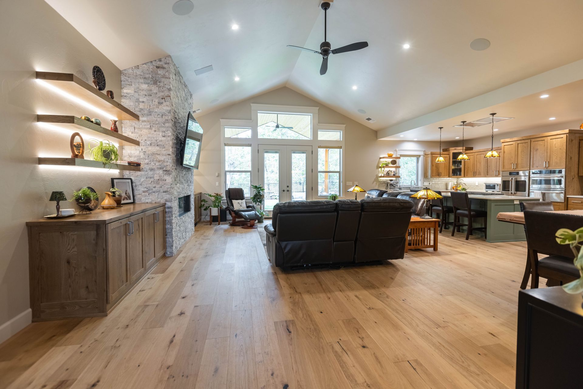 Open-concept living room and kitchen with wood floors, stone fireplace, black sofa, and vaulted ceiling