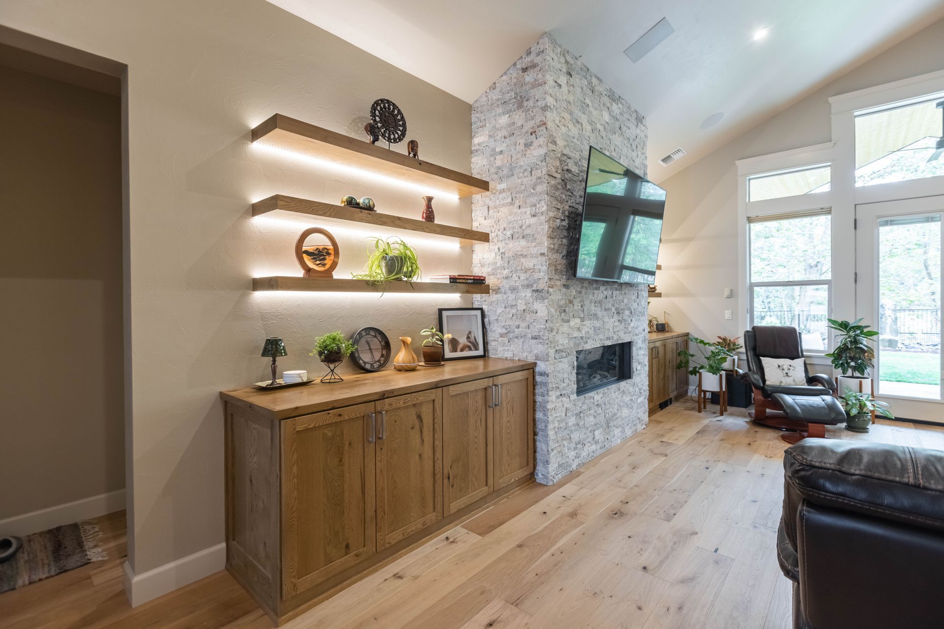 Modern living room with wood cabinet, floating shelves, stone fireplace, and large windows