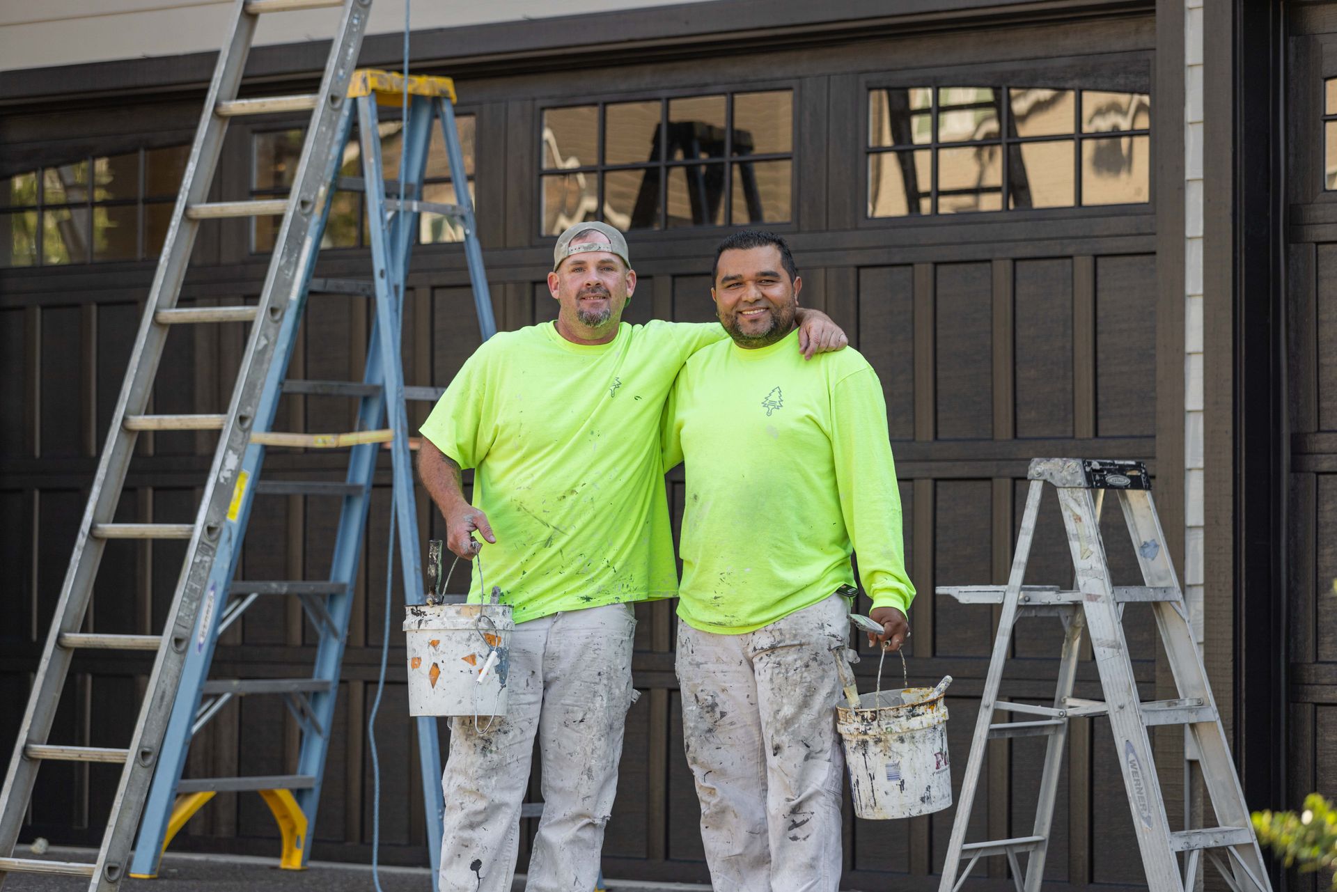 Two painters in neon shirts stand on a porch with ladders, holding paint buckets.