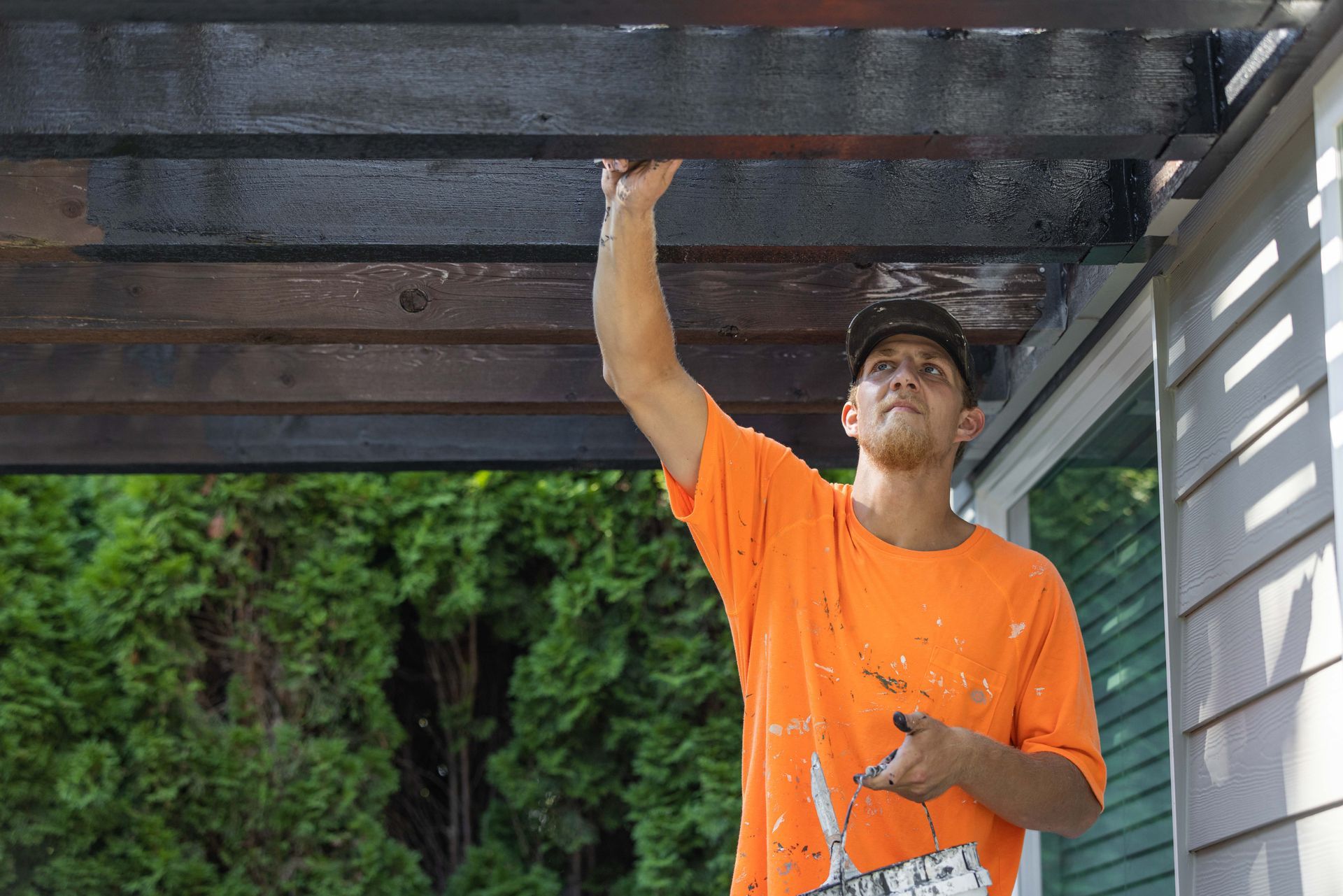 Man in orange shirt painting the underside of a porch roof outdoors