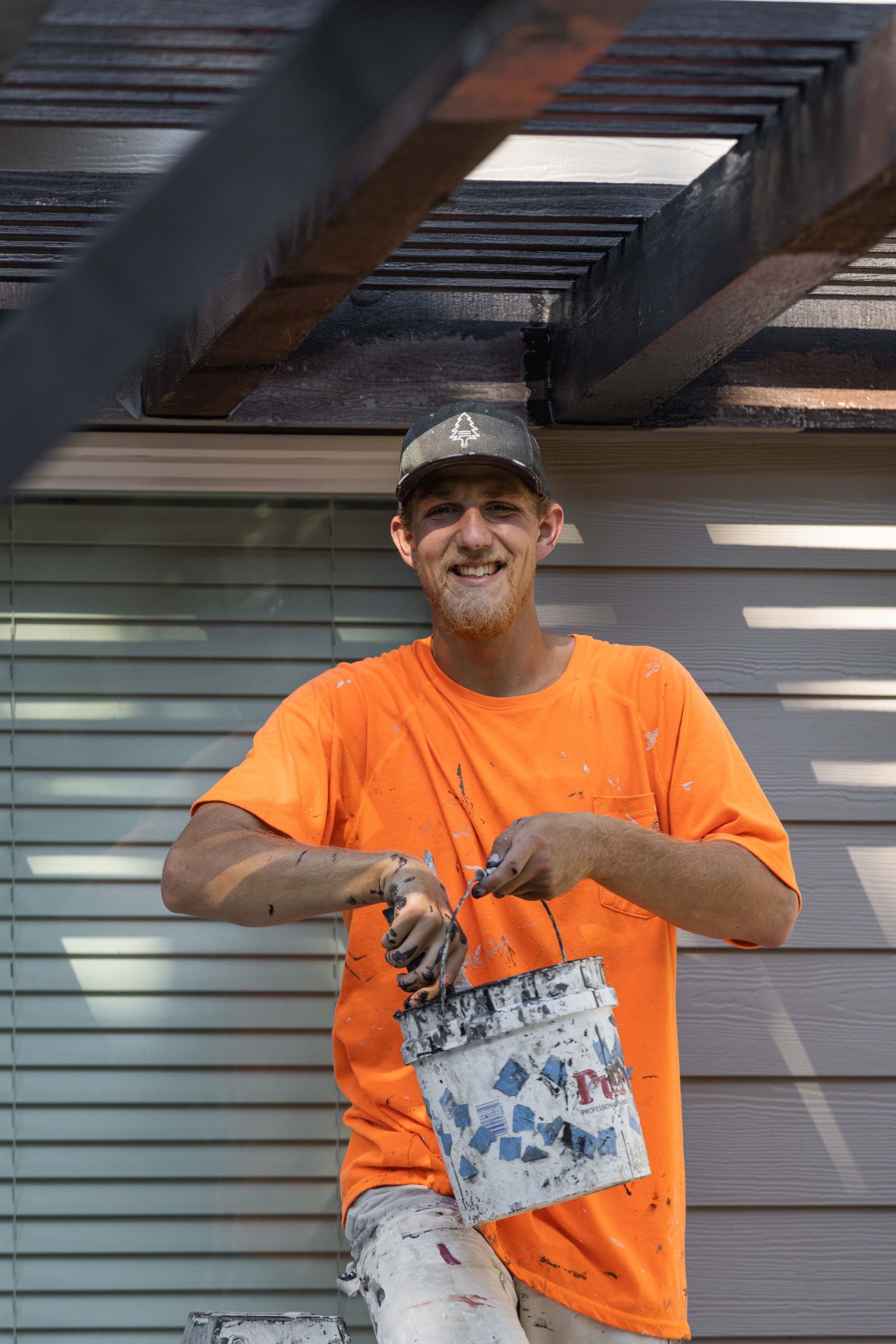 Man in bright orange shirt and cap holding a white bag, standing by a gray building exterior.