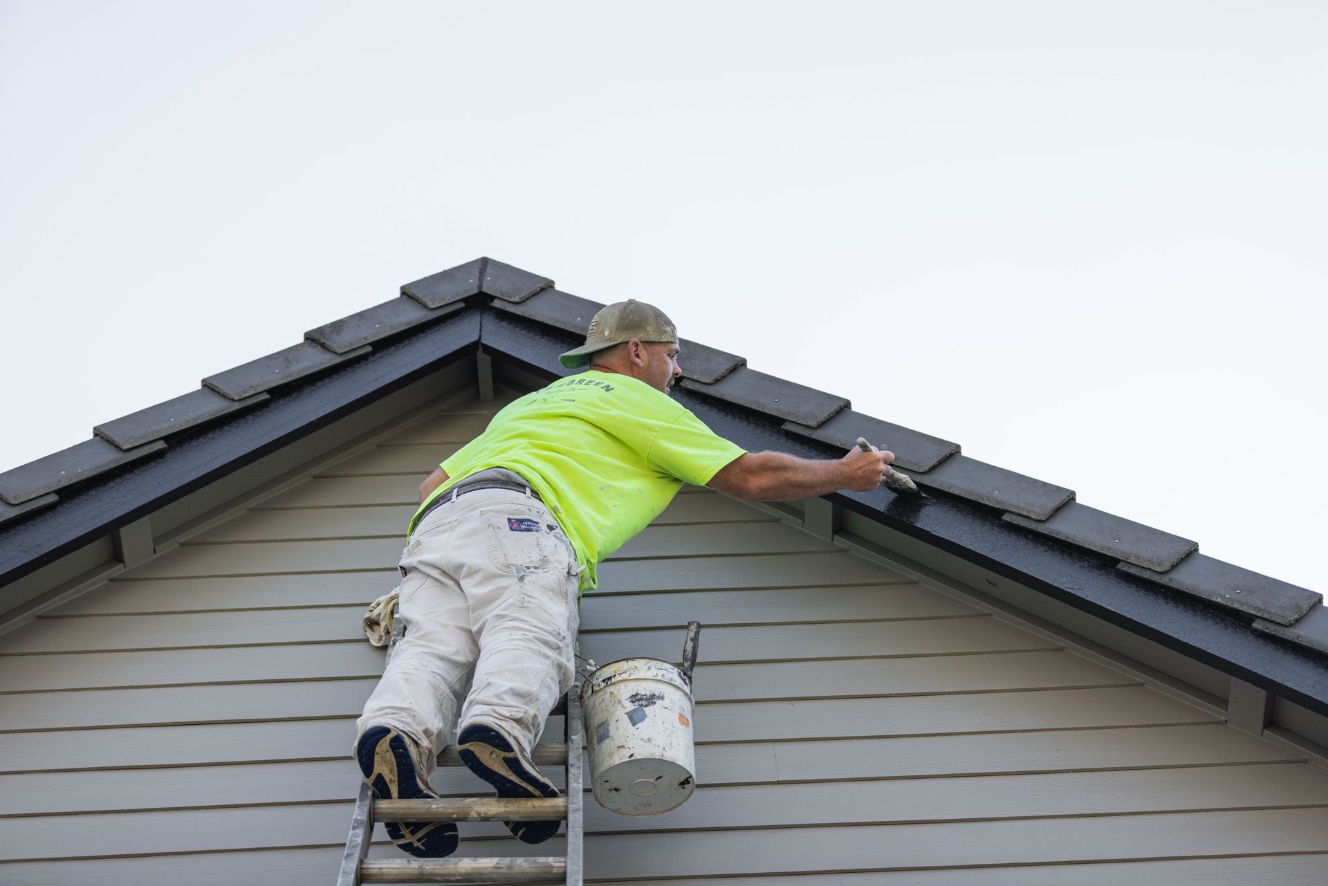 Worker painting house trim from ladder on roof peak, wearing neon shirt and white pants.