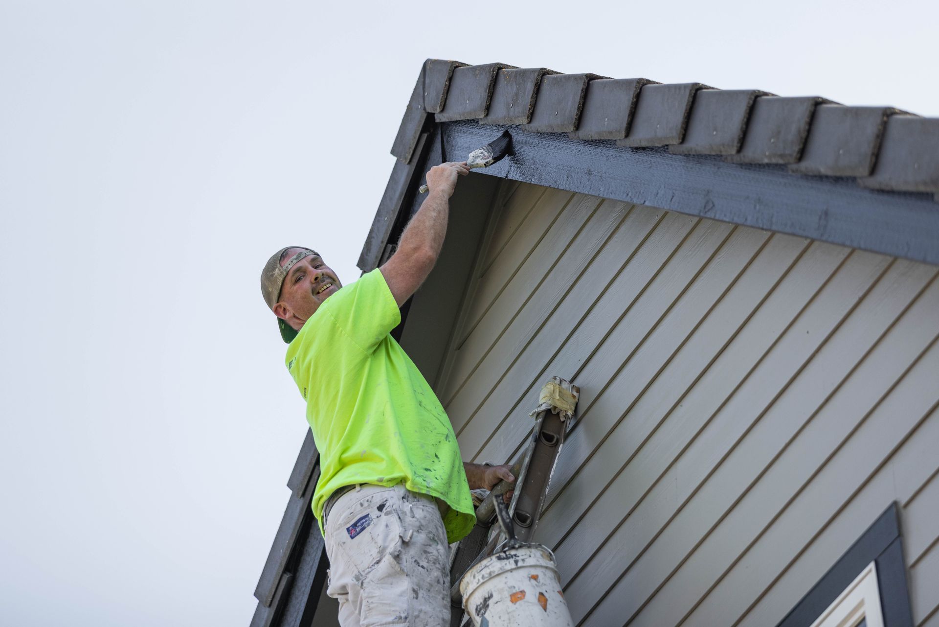 Worker in neon green shirt painting the eaves of a house near the roofline.