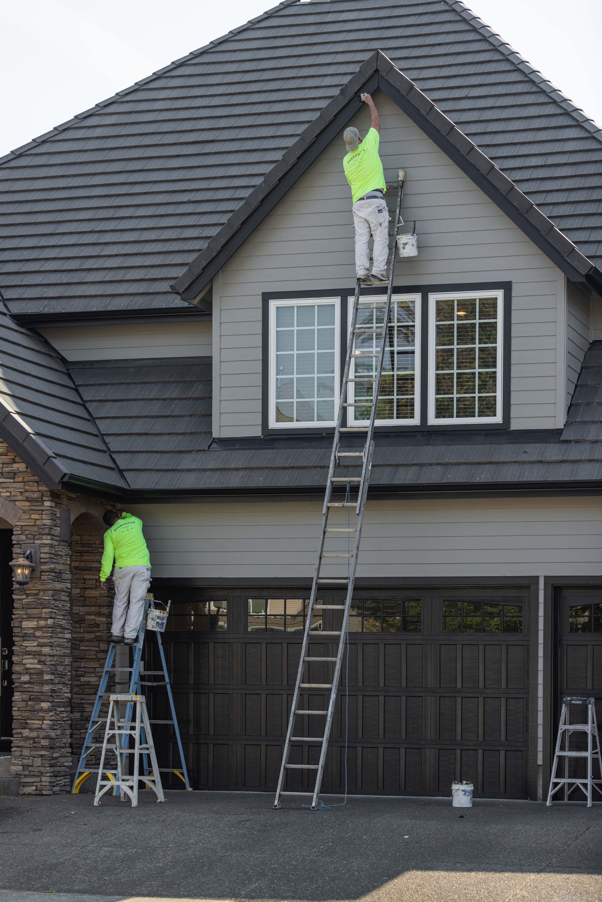 Two workers on ladders painting a gray house roof and upper front exterior.