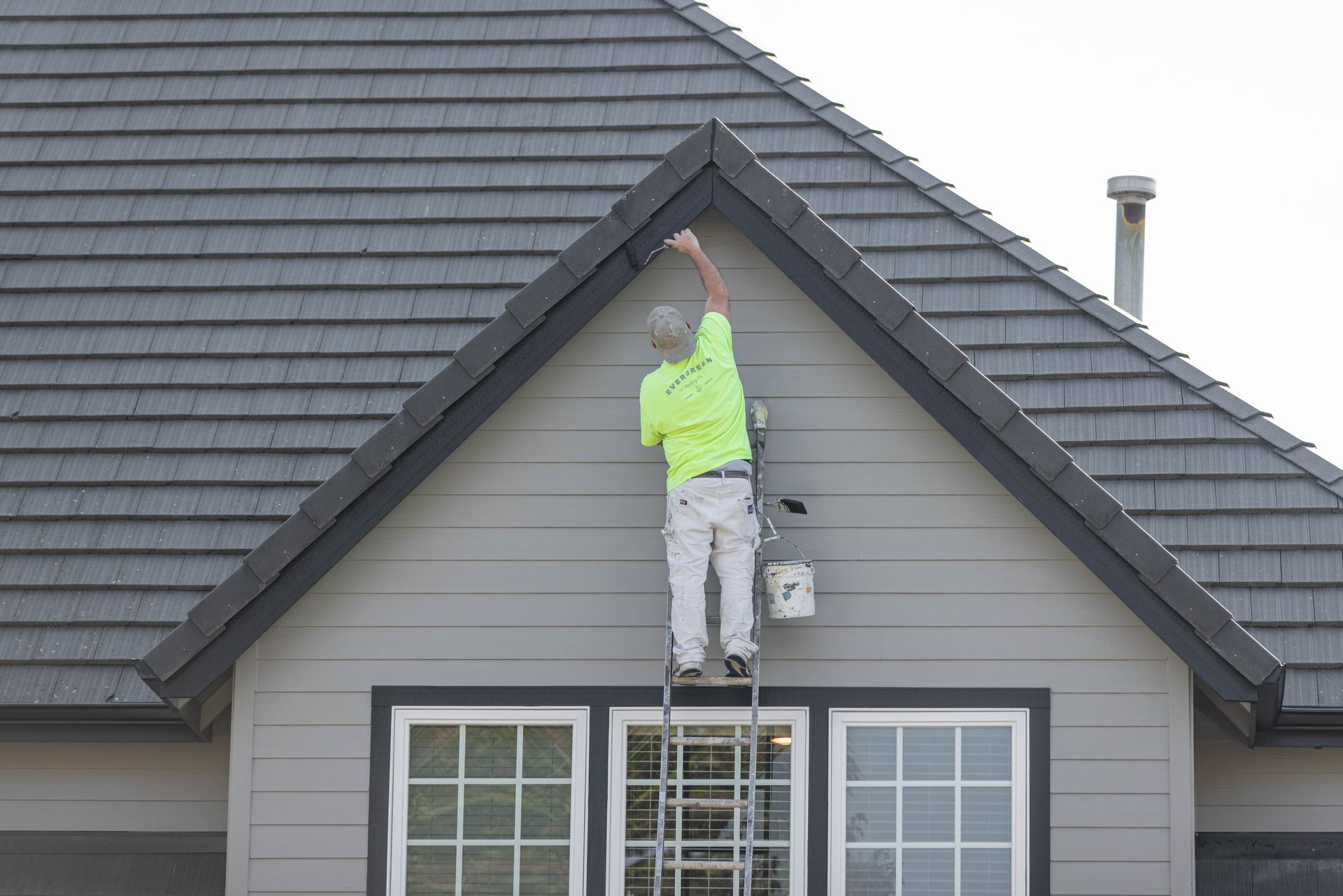 Worker in a neon shirt repairing the roof peak on a house above a ladder.