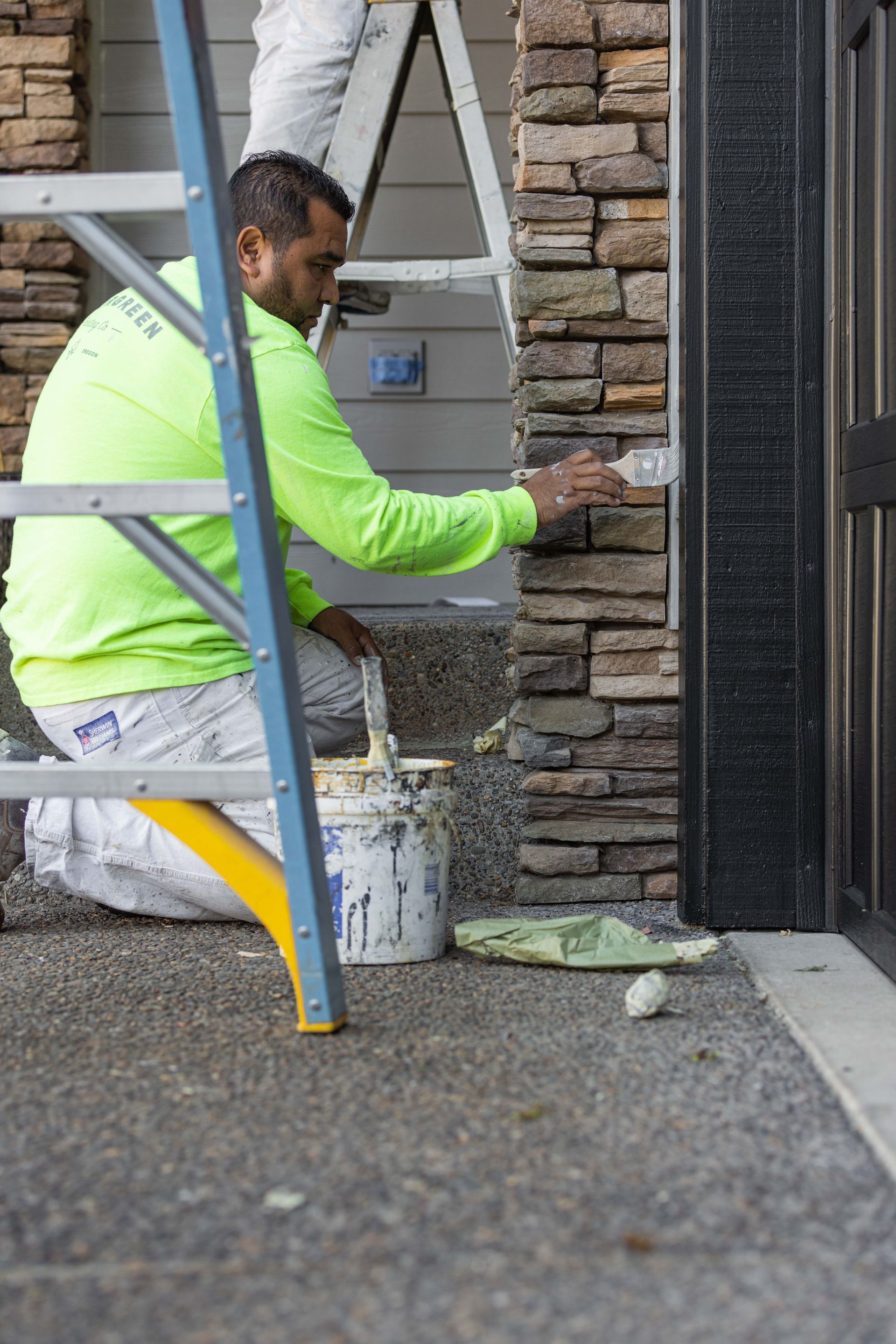 Worker in a neon jacket applying mortar to a stone wall beside a ladder.