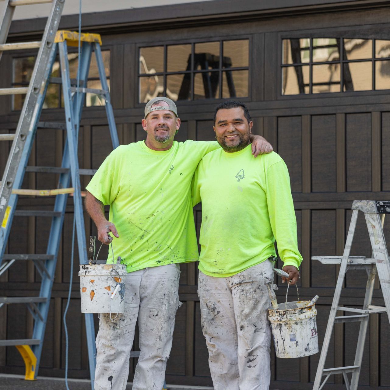 Two painters in neon green shirts stand in front of a house, holding paint buckets and smiling.