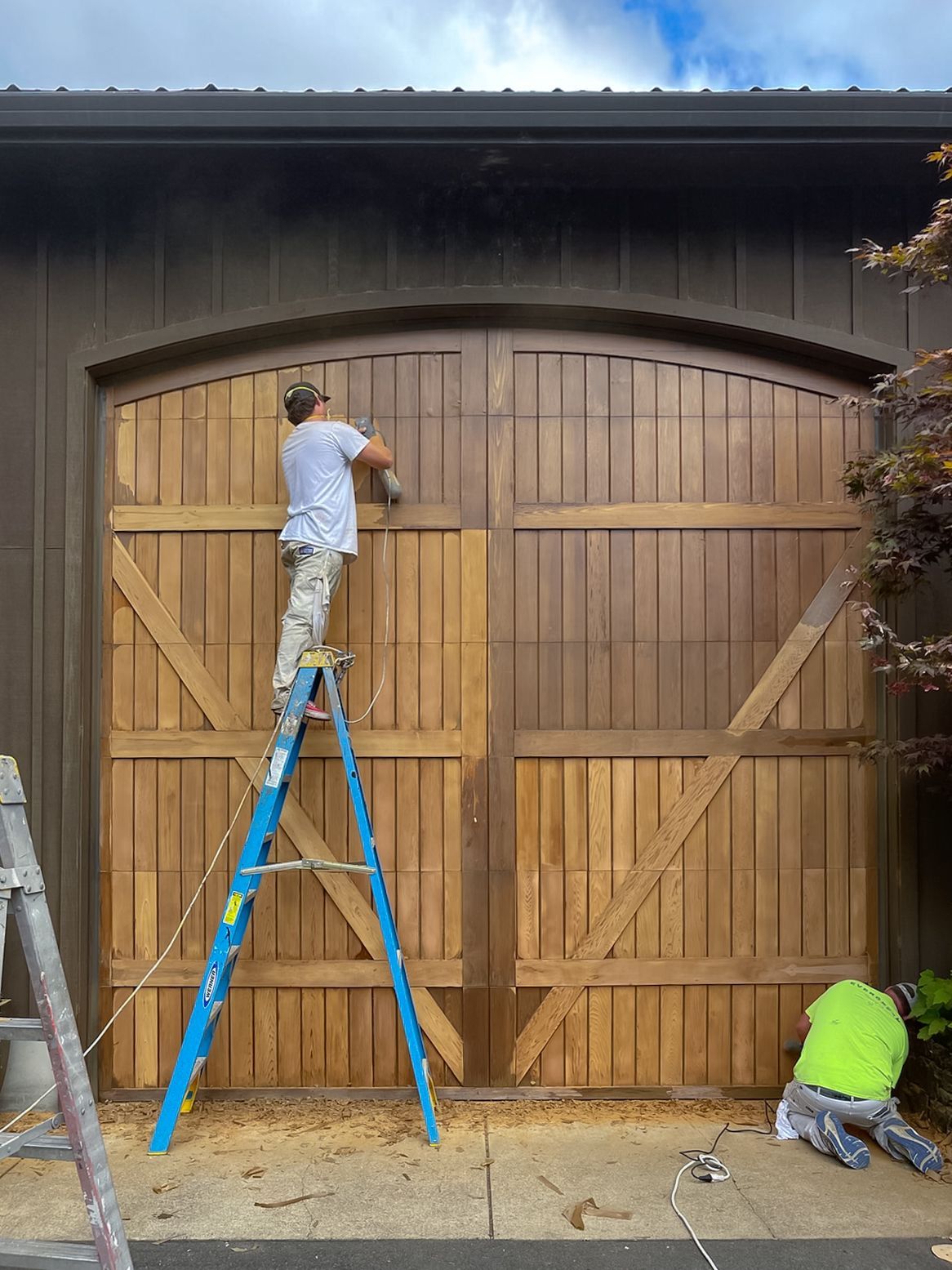 Workers on ladders painting a large wooden garage door under a dark roof overhang