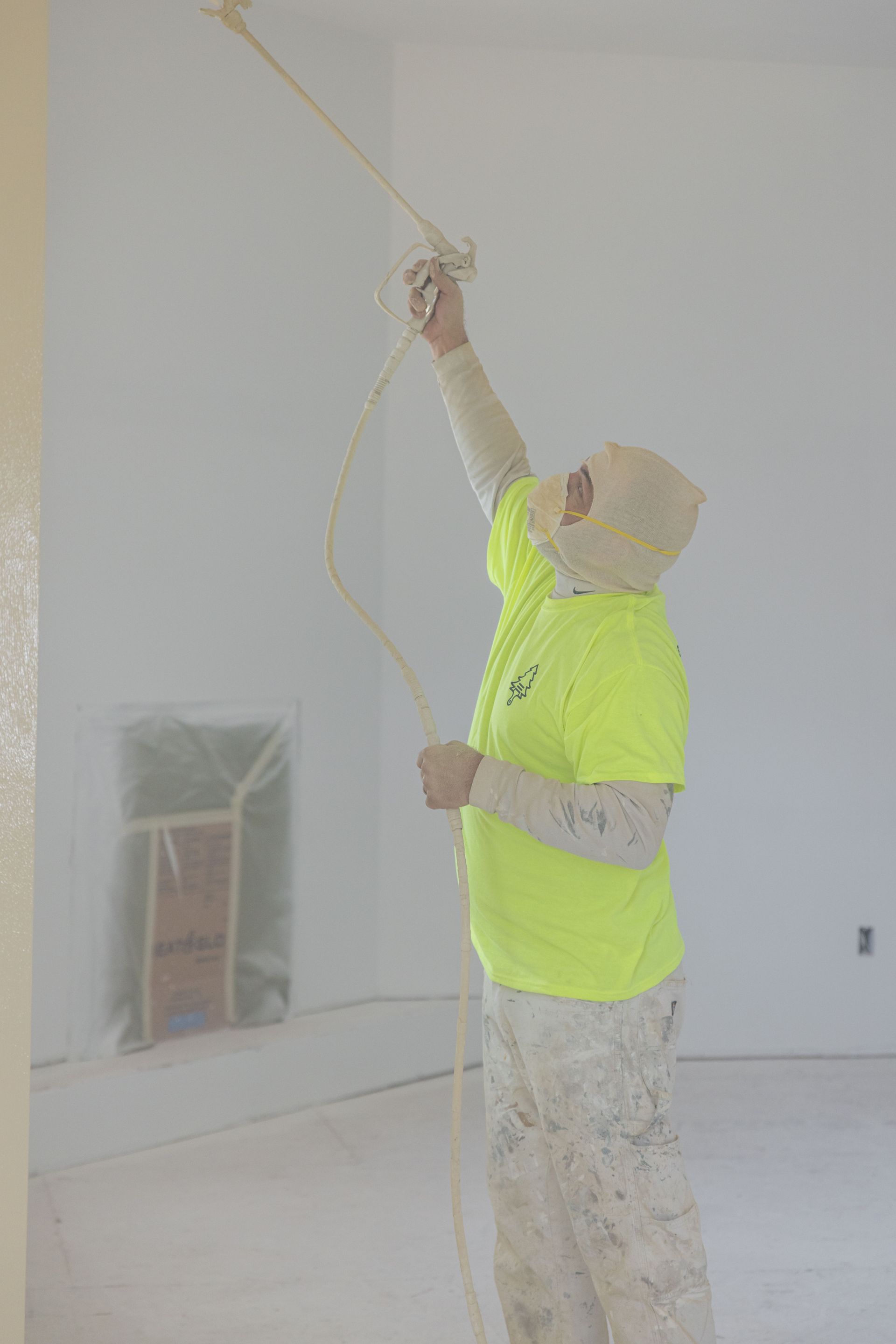 Worker in a bright yellow shirt painting a ceiling with a roller in a room under renovation