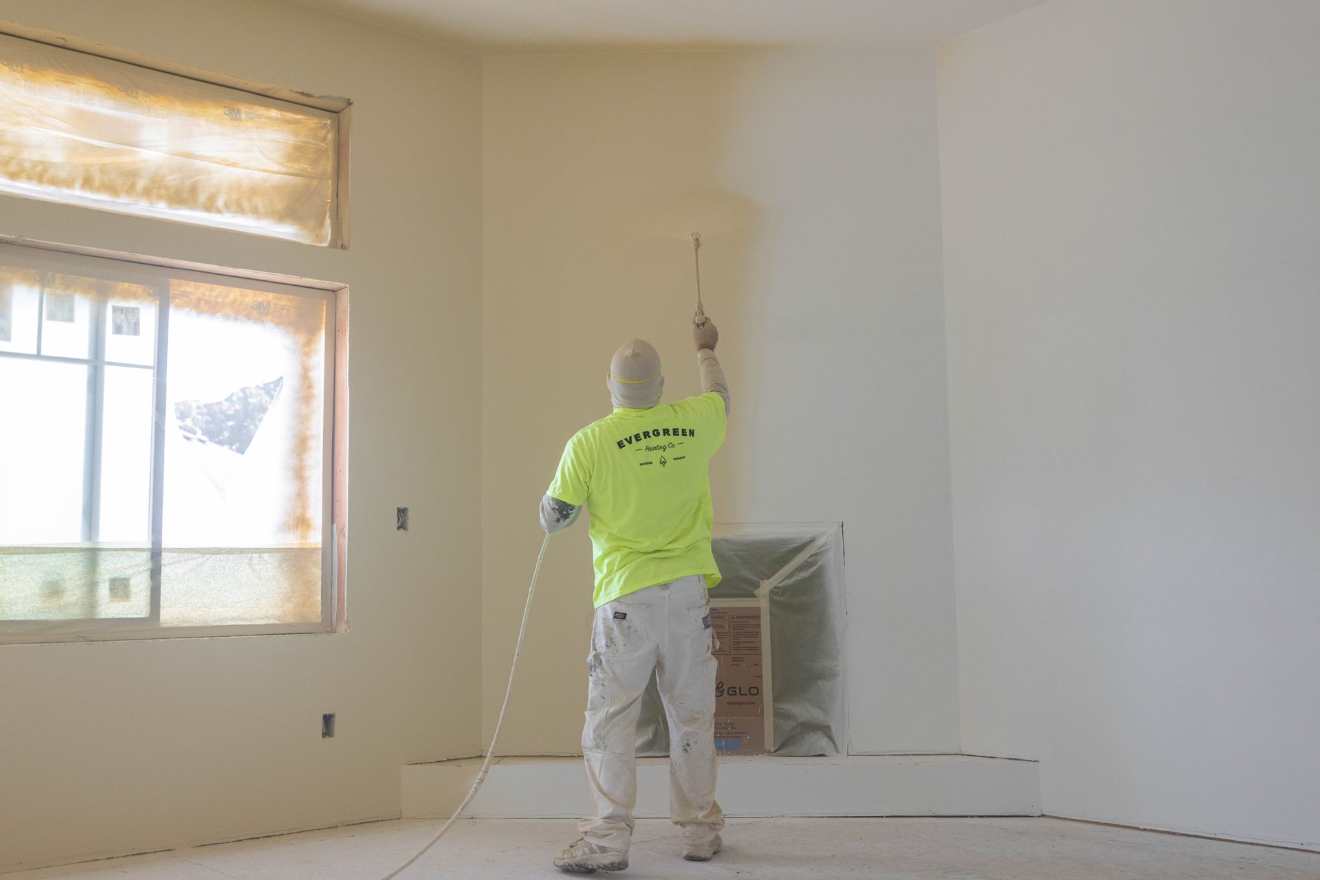 Worker painting a white wall in a bright room with a window and covered fireplace.