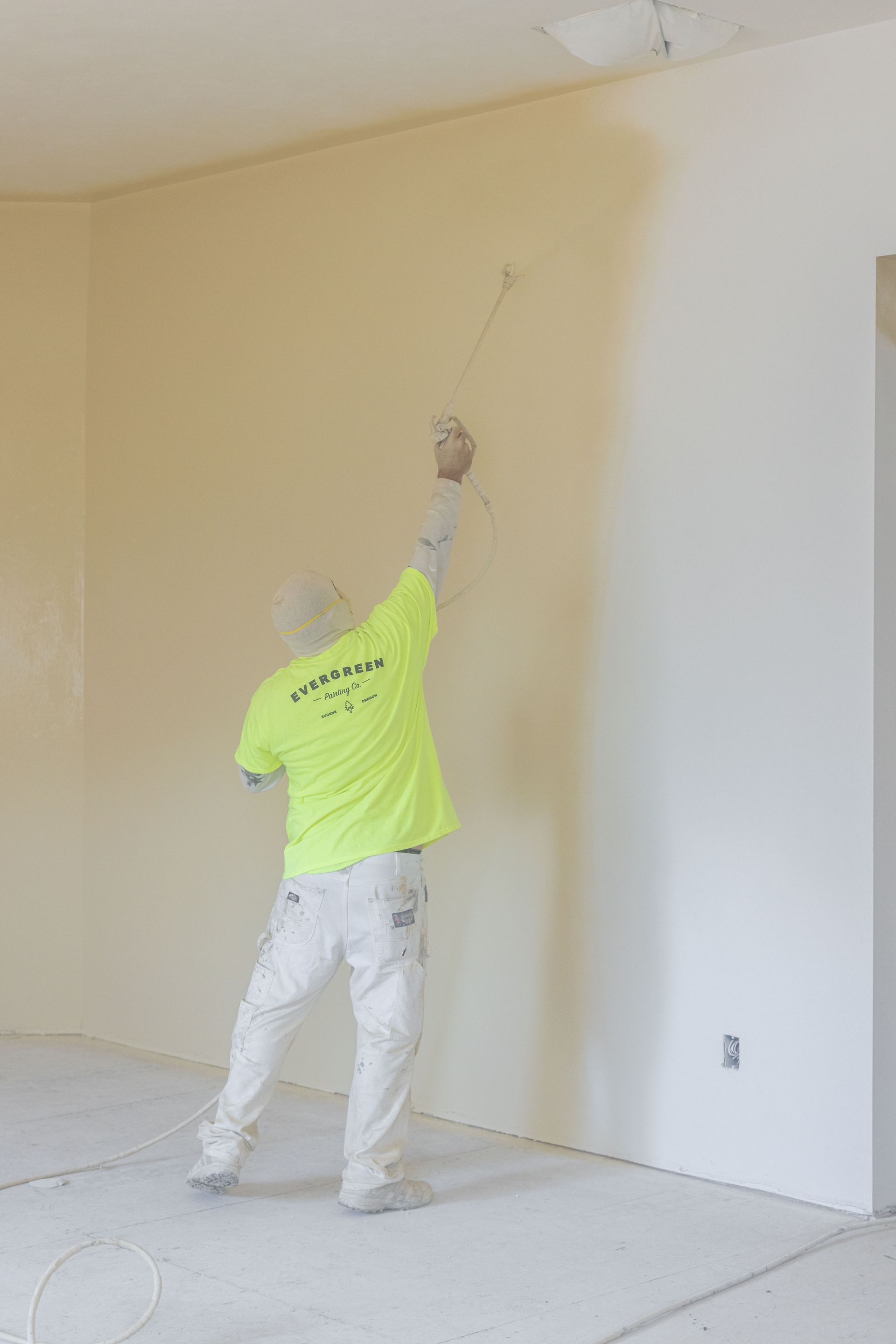 Person in a neon shirt painting a beige wall in an unfinished room.