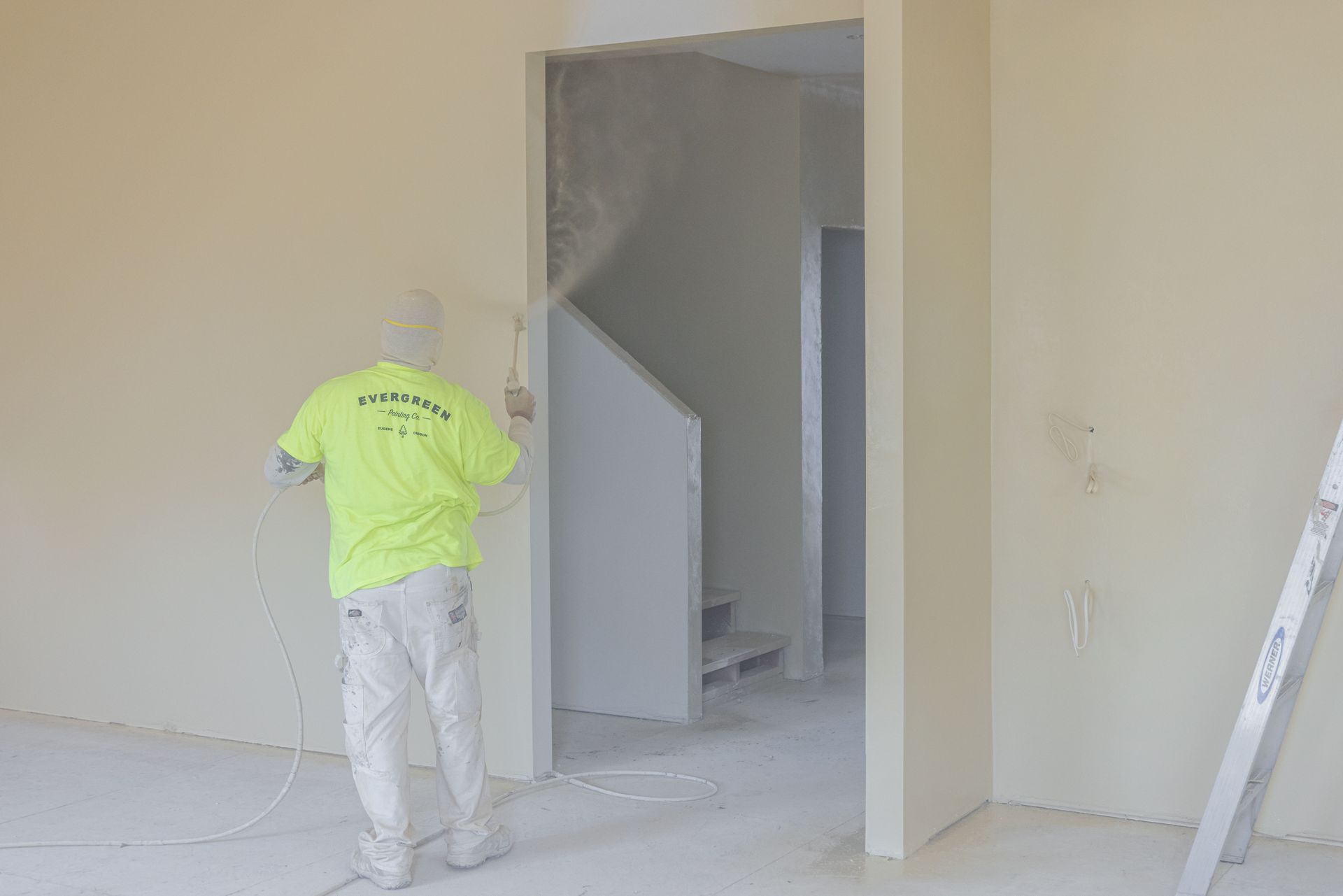 Worker in neon shirt painting a beige interior wall near a doorway.