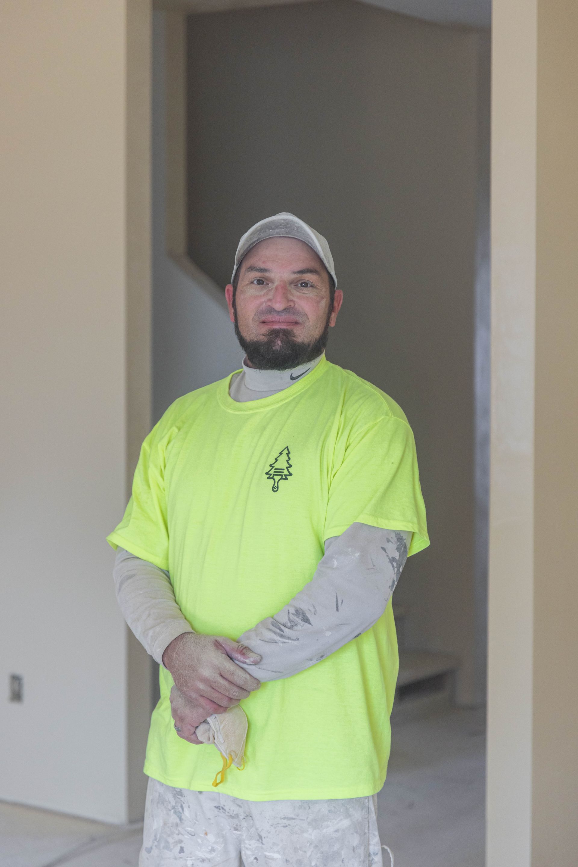 Man standing indoors in a neon yellow shirt and gray cap, hands clasped, in a doorway.