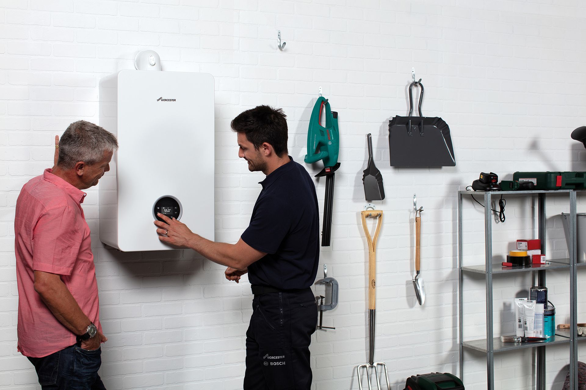 Two men are standing next to each other in a garage looking at a boiler.