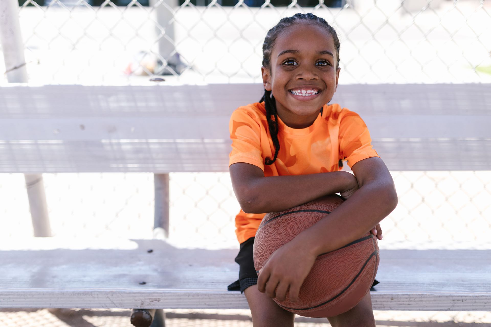 Smiling child in an orange shirt holding a basketball on a white bench outdoors