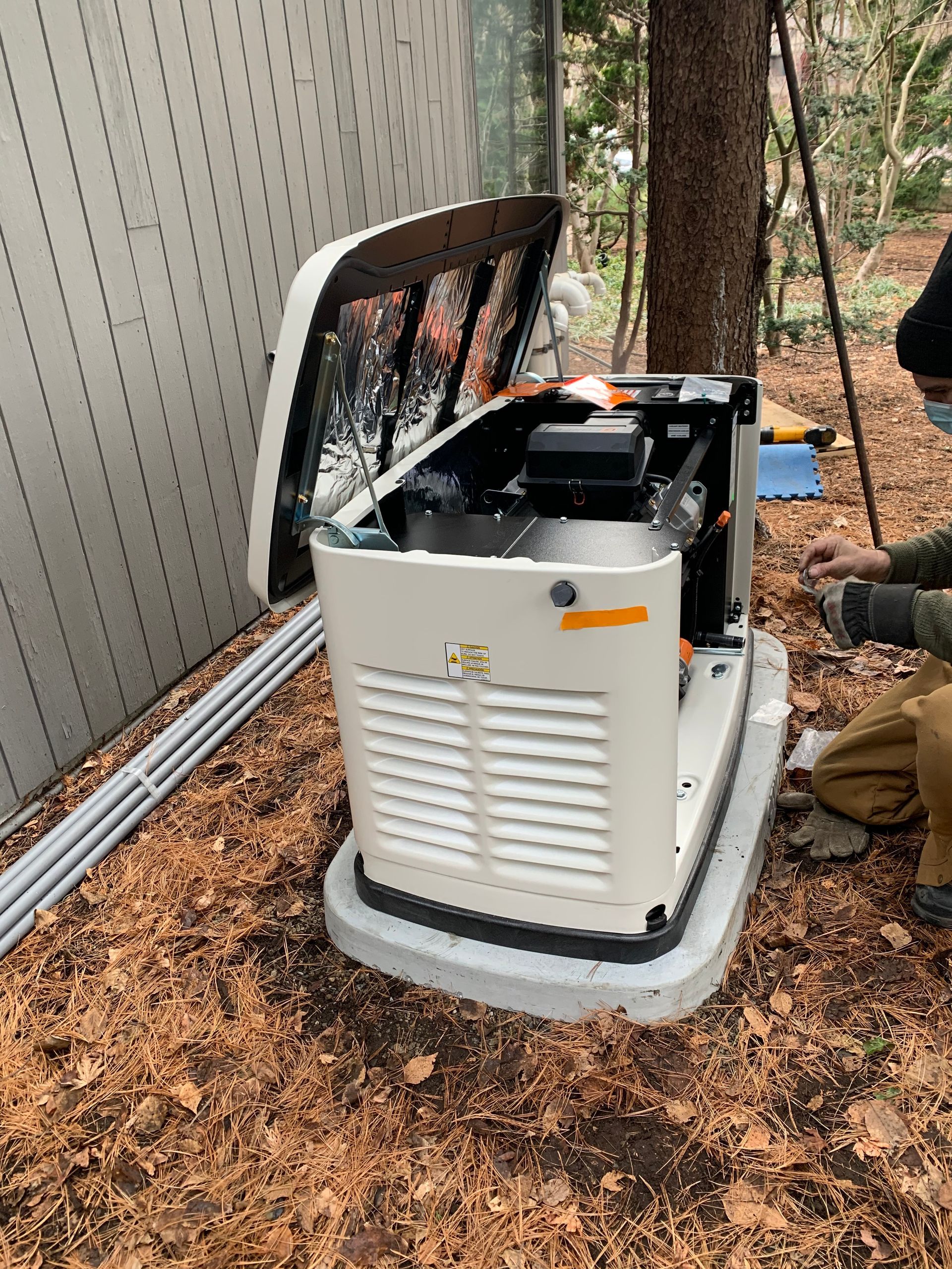 Generator with open access panel, being serviced by a person outdoors near a building and trees.