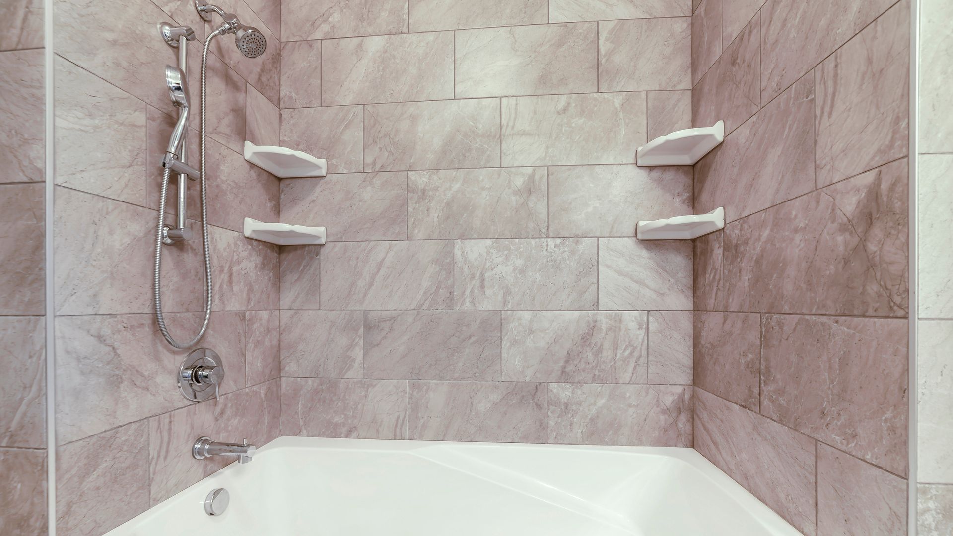 Pink-tiled bathroom shower and bathtub with wall shelves and handheld showerhead Pink-tiled bathroom shower and bathtub with wall shelves and handheld showerhead