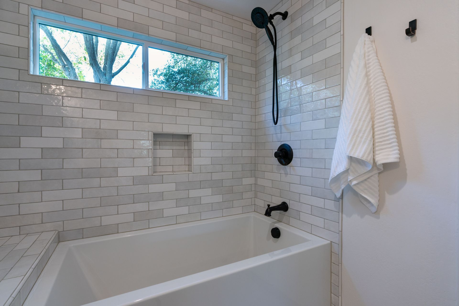 Modern white-tiled bathroom with a bathtub, shower fixtures, window, and hanging towel Modern white-tiled bathroom with a bathtub, shower fixtures, window, and hanging towel