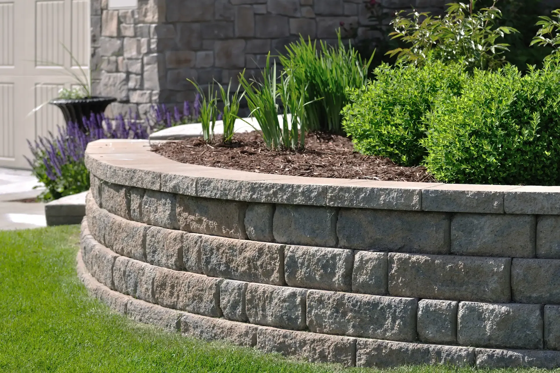Curved retaining wall made of gray bricks holds a garden bed with plants, next to a grassy lawn.
