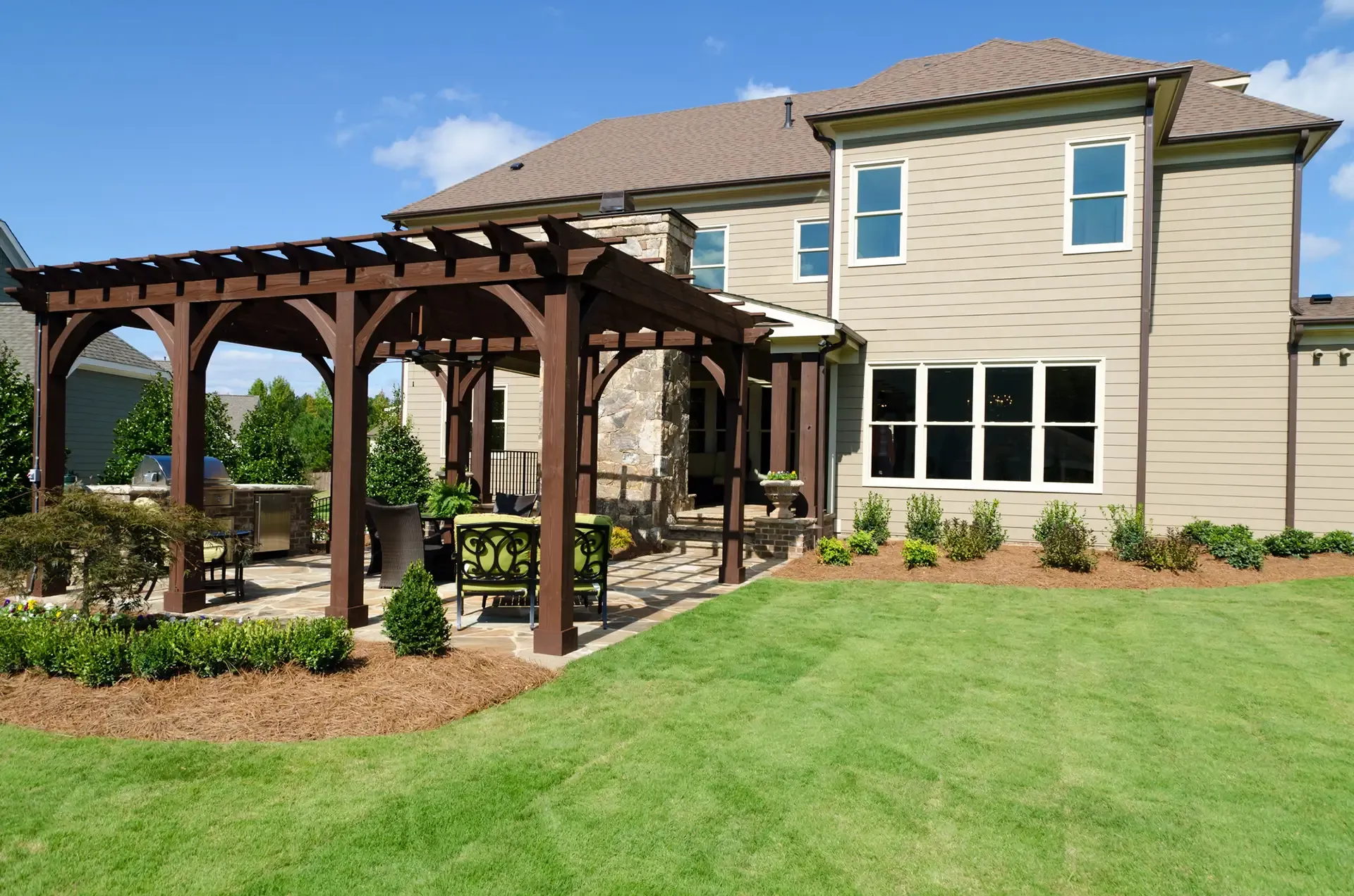 Backyard with brown pergola over a patio, connecting to a two-story house with green lawn and landscaping.