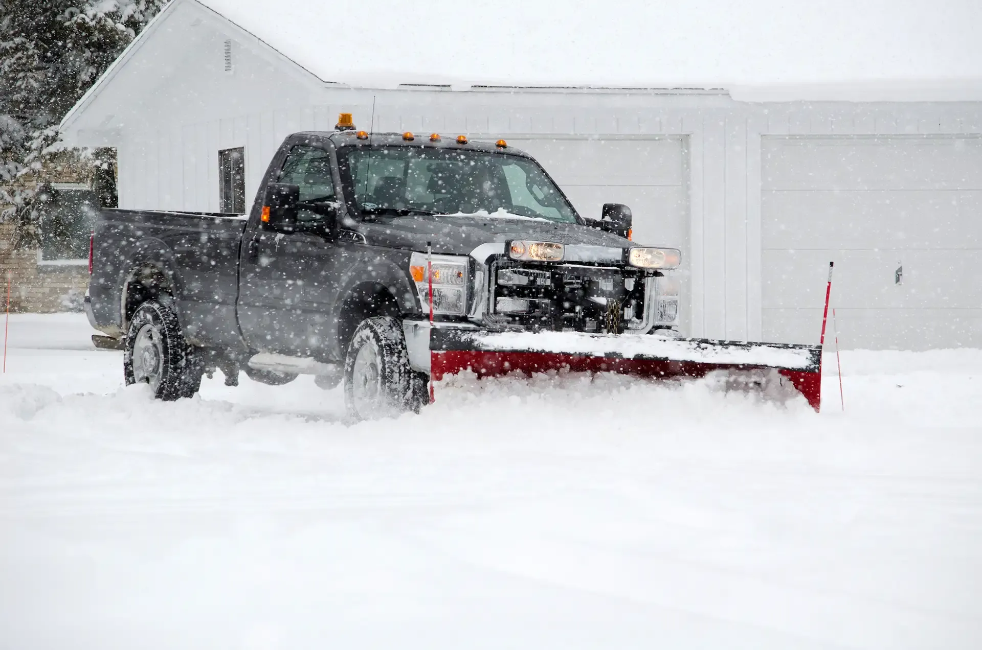 Black pickup truck with snowplow clearing snow in front of a white house during a snowstorm.