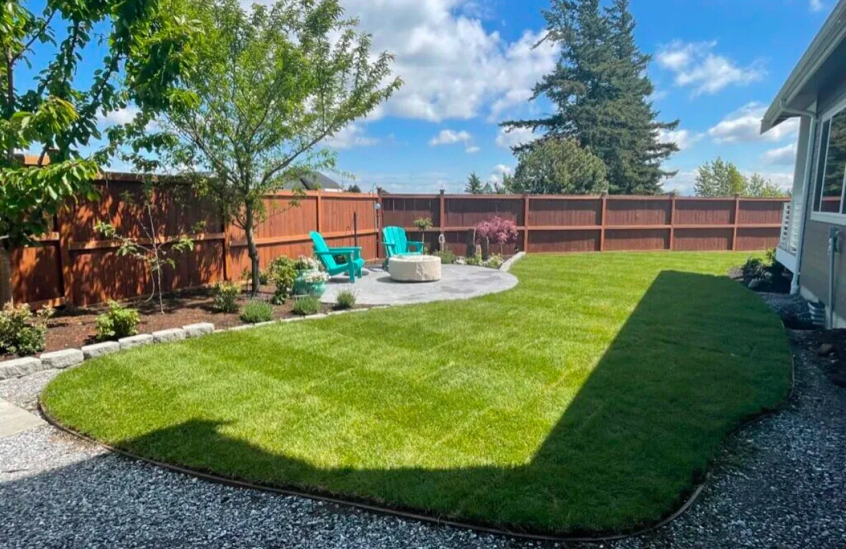 Backyard with a freshly mowed green lawn, stone fire pit, wooden fence, two blue chairs, and gravel path.