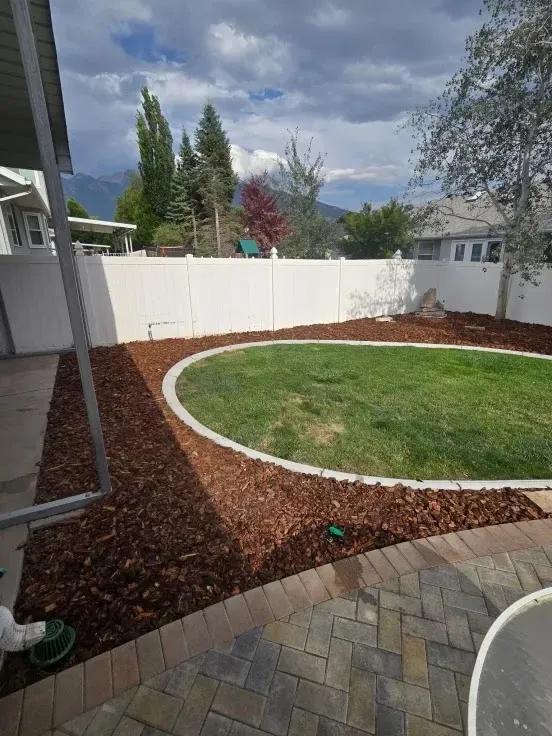 Backyard with a lawn edged by a curved concrete border, surrounded by wood mulch. White fence and cloudy sky.