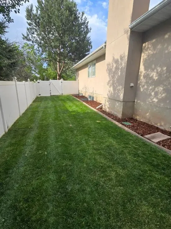 Green lawn next to a white fence and tan house with mulch border under a partly cloudy sky.