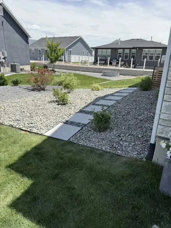 Backyard with stone path, gravel beds, lawn, and several houses under a cloudy sky.