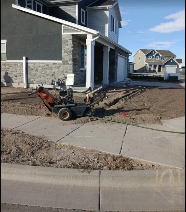 A trench is being dug near a sidewalk and house by a machine. Brown dirt and green grass visible.