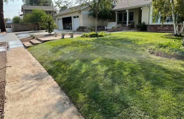 Lawn sprinklers watering a green lawn in front of a beige house with a walkway and street in the foreground.
