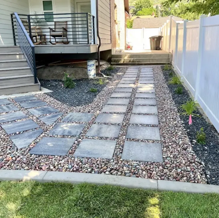 Stone pathway with square pavers and gravel beside a house with a deck and stairs, next to a white fence.