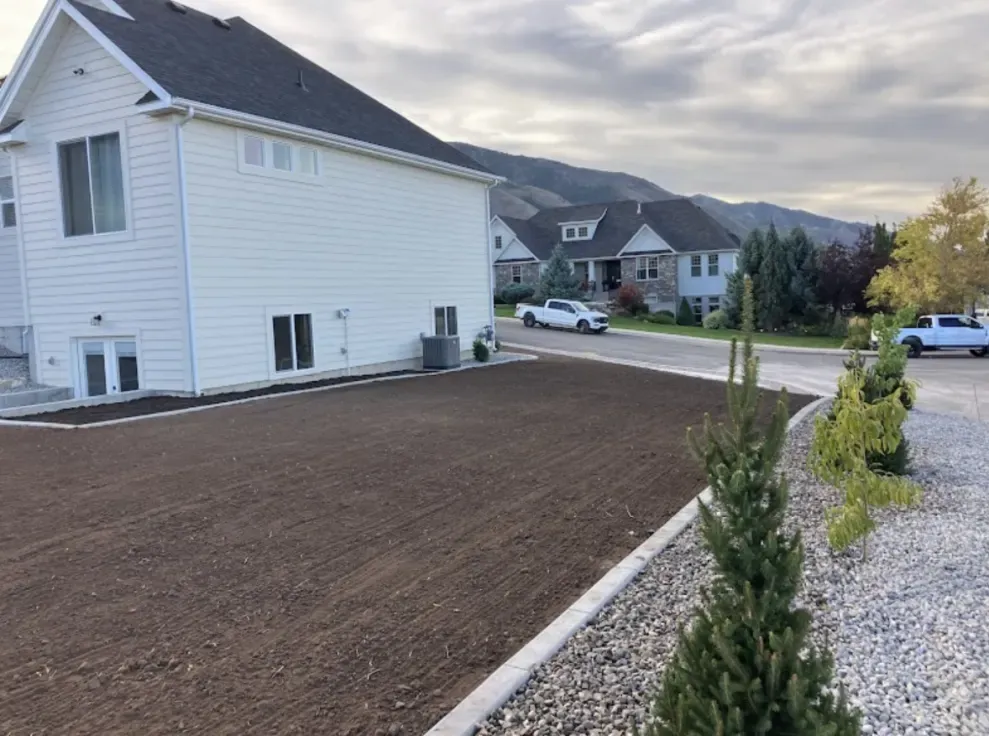 Freshly tilled yard next to a white house, street, and mountains under a cloudy sky.