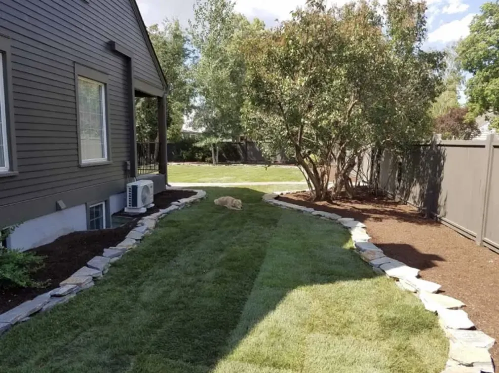 Side yard with fresh green grass, stone edging, dark house, and a brown fence.