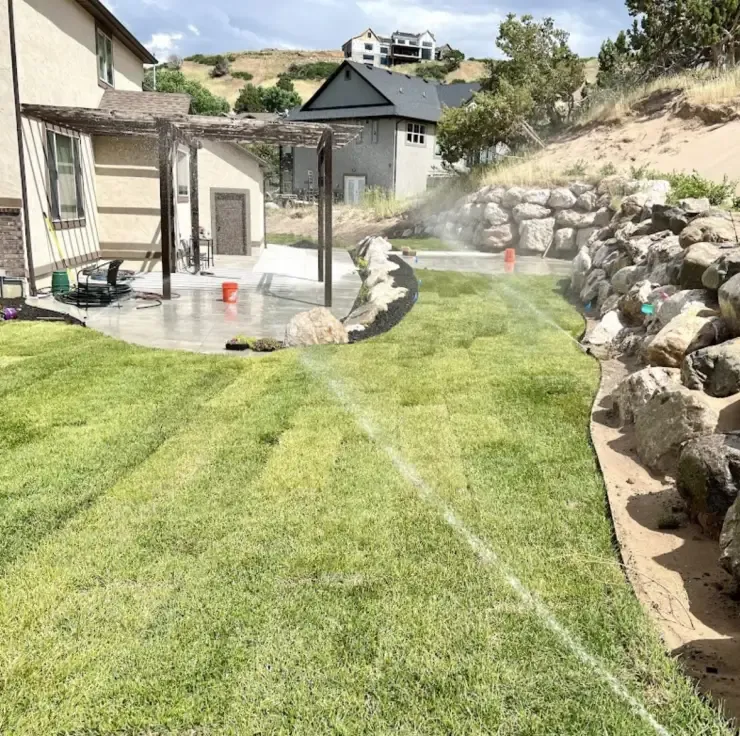 Backyard with green grass, stone retaining wall, and patio; sprinkler watering the lawn.