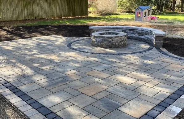 Stone patio with fire pit, surrounded by a low wall, in a backyard setting.