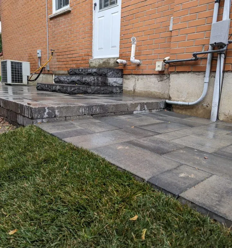 Stone patio and steps leading to a white door of a brick building; grass in foreground.