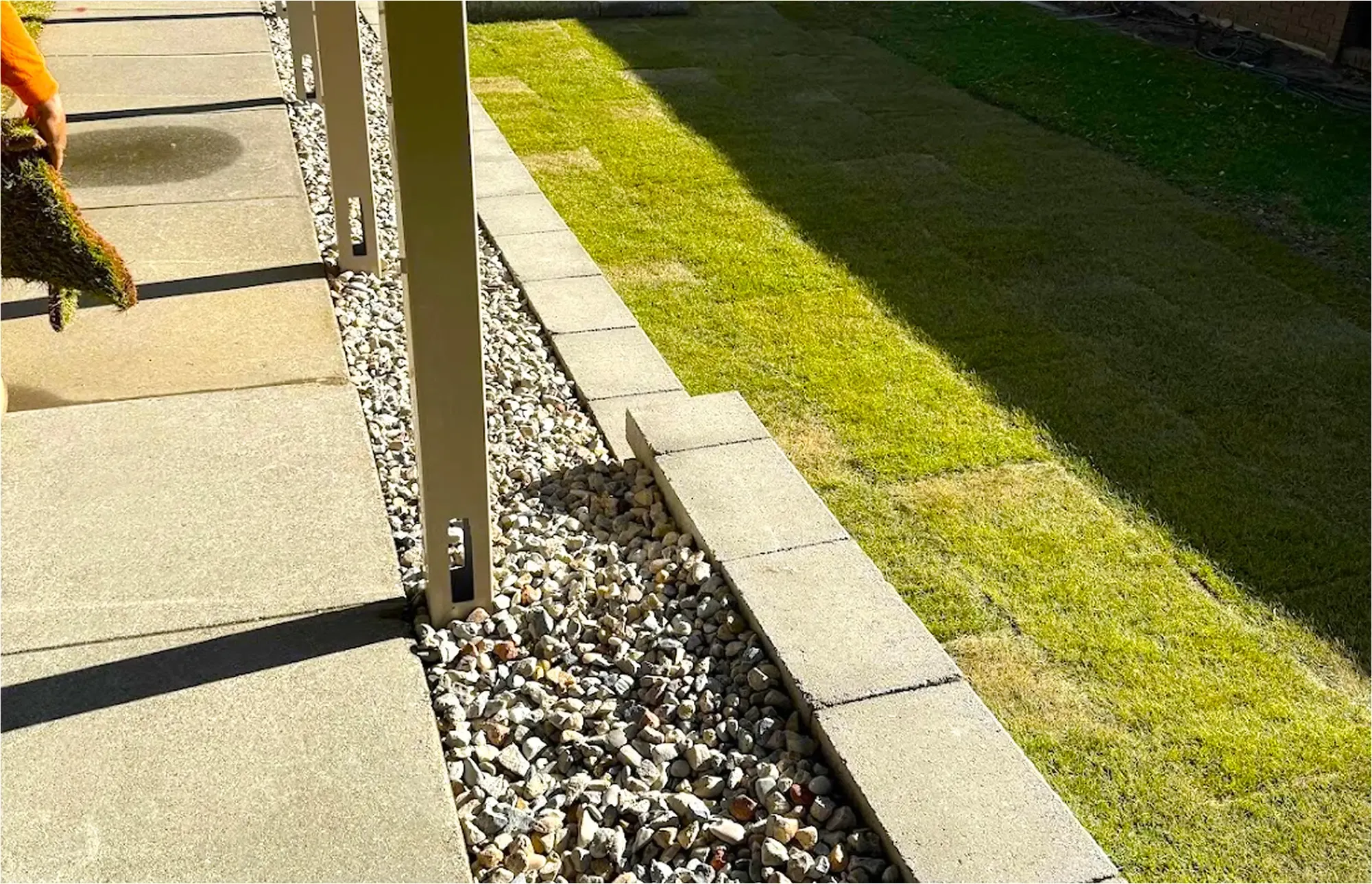 Stone pavers border a gravel bed, concrete blocks, and a green lawn under sunlight.