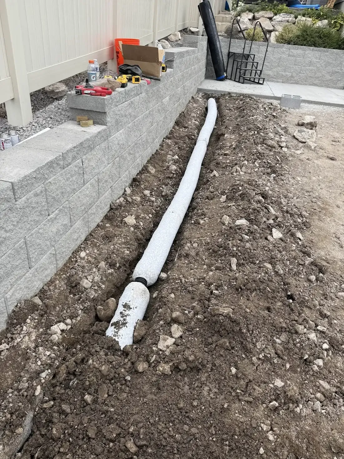 Drainage system installation: white sock-like pipe in a trench, alongside a stone wall and fence.