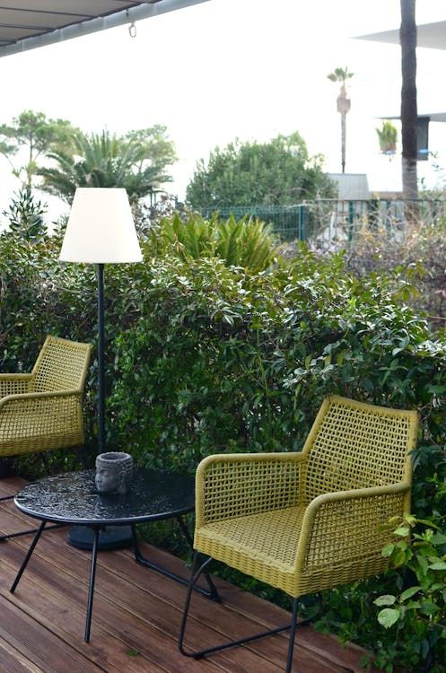 Two green woven chairs and a table on a wooden deck with a tall lamp and lush greenery. Two green woven chairs and a table on a wooden deck with a tall lamp and lush greenery.