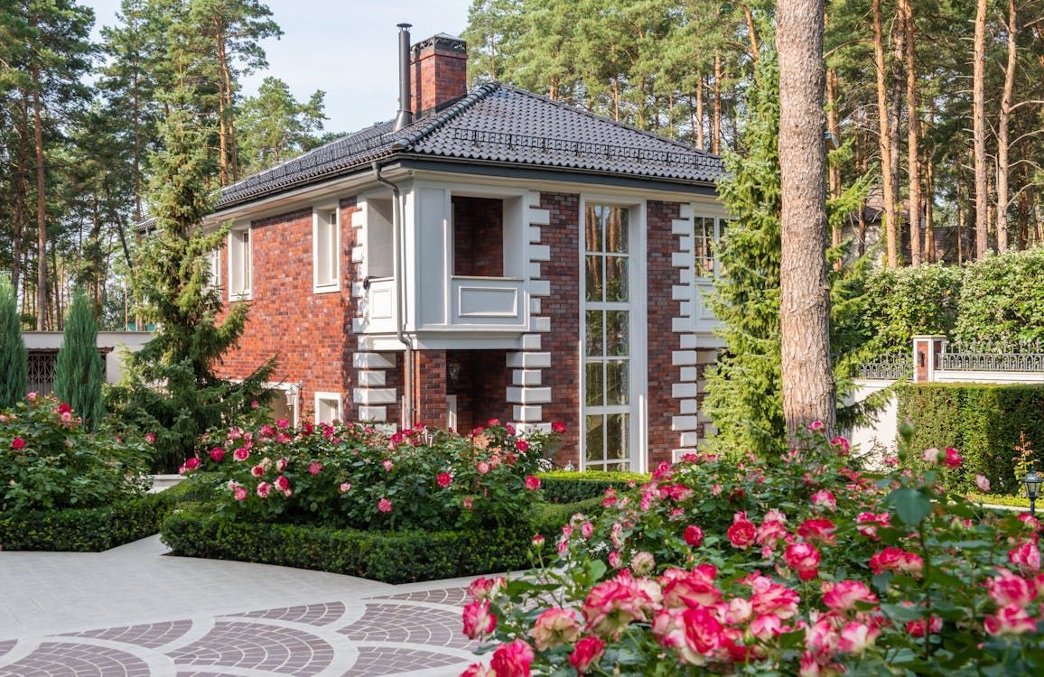 Brick house with white trim, surrounded by rose bushes, on a paved driveway in a wooded area. Brick house with white trim, surrounded by rose bushes, on a paved driveway in a wooded area.