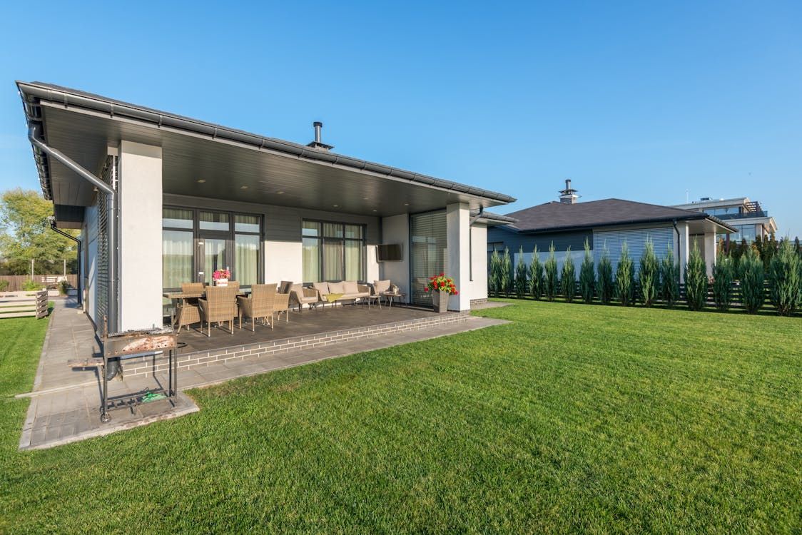 Modern house with covered patio, outdoor dining set, and green lawn on a sunny day. Modern house with covered patio, outdoor dining set, and green lawn on a sunny day.