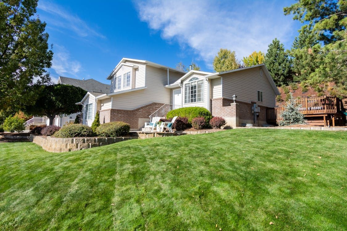 Two-story house with beige siding and brick base, on a grassy hill under a blue sky. Two-story house with beige siding and brick base, on a grassy hill under a blue sky.