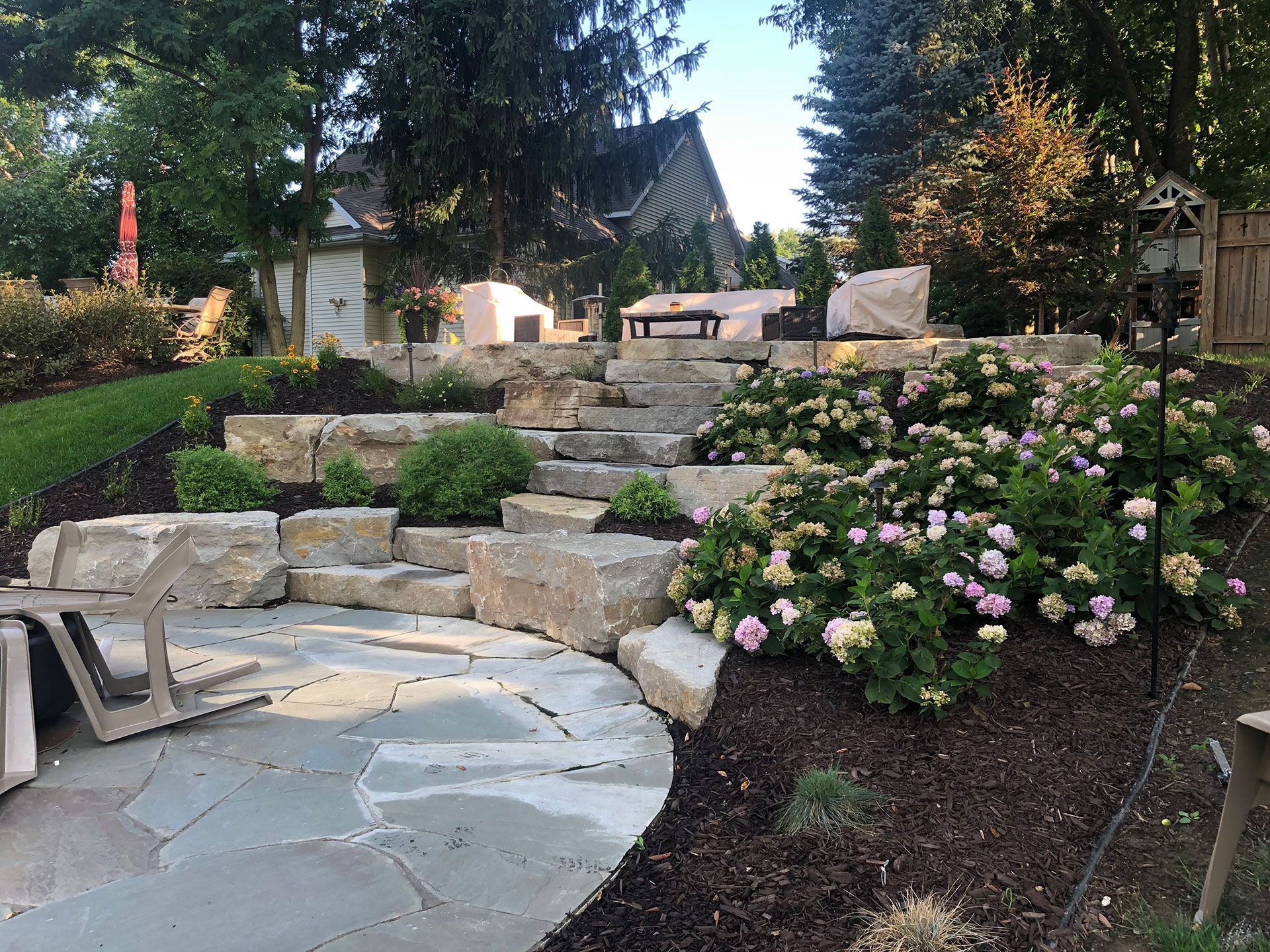 Stone patio and stairs lead to a seating area in a landscaped backyard with flowering bushes. Stone patio and stairs lead to a seating area in a landscaped backyard with flowering bushes.