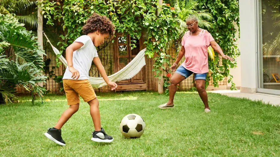 A child kicking a soccer ball while playing with an older person in a grassy backyard. A child kicking a soccer ball while playing with an older person in a grassy backyard.
