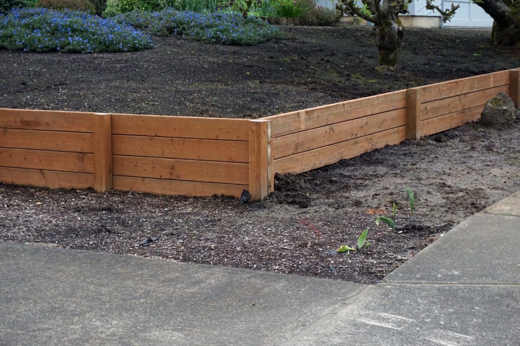 Brown wooden retaining wall bordering a flower bed and a sidewalk.