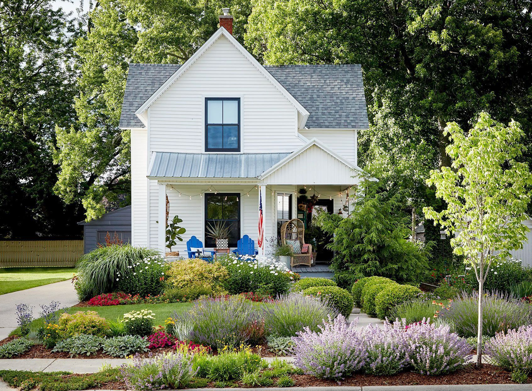 White two-story house with a vibrant garden; blue chairs on porch, lush greenery, and a bright sunny day. White two-story house with a vibrant garden; blue chairs on porch, lush greenery, and a bright sunny day.