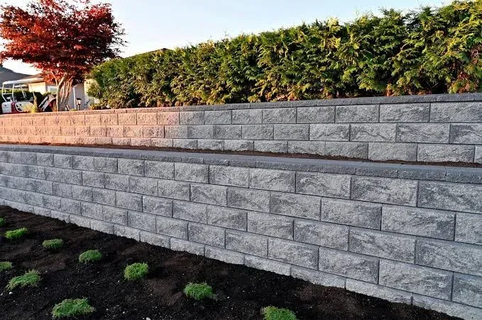 Two-tiered gray brick retaining wall with small bushes in front and green hedge behind.