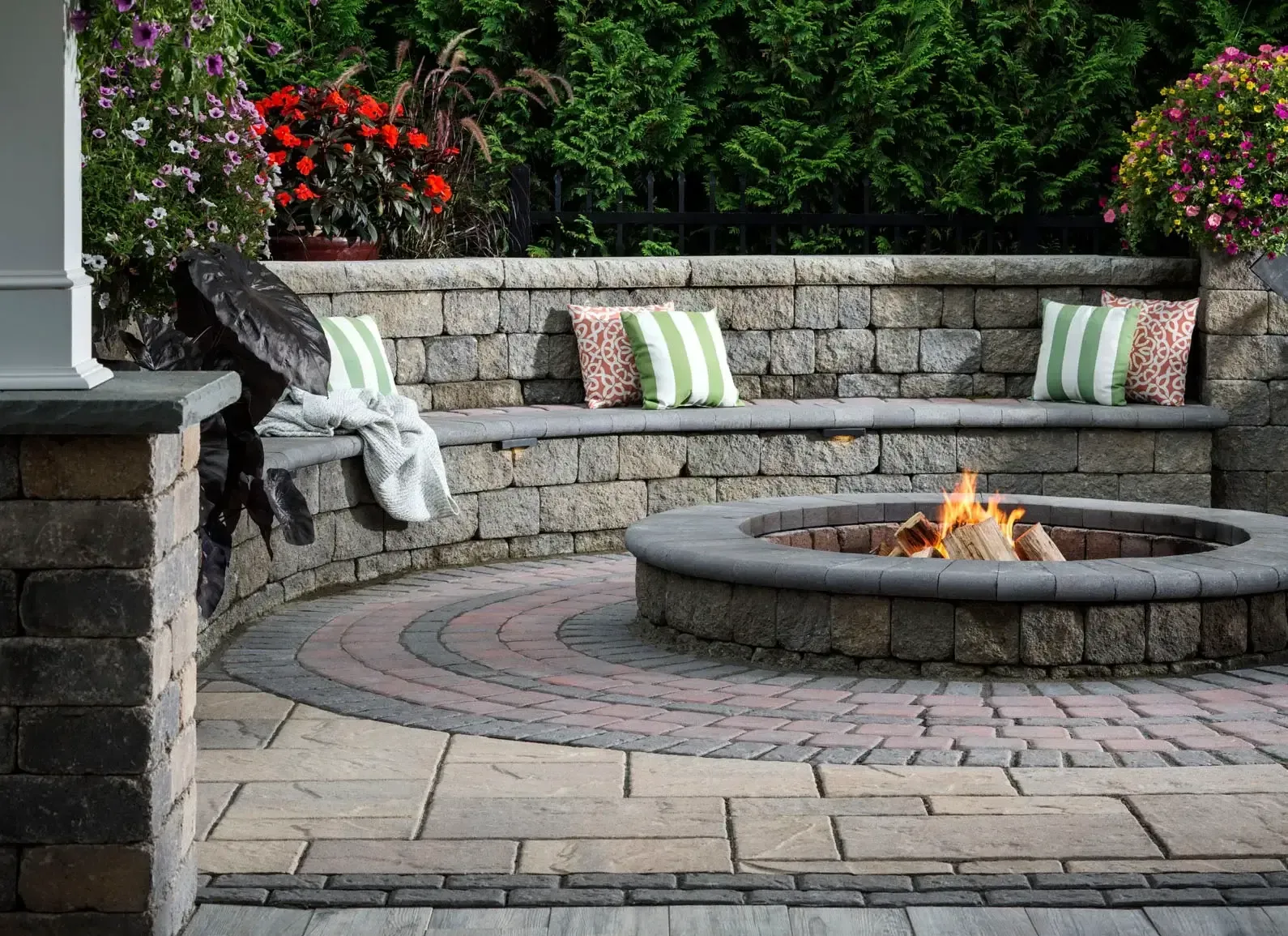 Stone patio with built-in bench and fire pit, green and pink patterned pillows, flowers, and a blanket.
